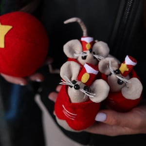 May include: A red felt ball with a yellow star and several handmade mouse figurines. The mice have beige bodies, red outfits, and small hats. The figurines have black bead eyes and white whiskers. The theme is circus or carnival.