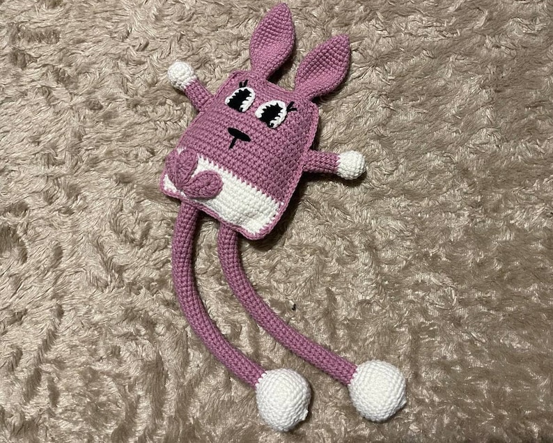 May include: A pink crocheted bunny with long legs and white feet. The bunny has large black eyes and a white nose.