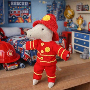 Puede incluir: Un ratón de peluche vestido con un uniforme de bombero rojo con detalles amarillos y un sombrero a juego. El juguete está en una habitación infantil decorada con un tema de camión de bomberos, incluyendo una colcha y arte mural.