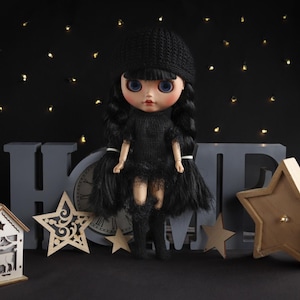 May include: A black doll with blue eyes and black hair wearing a black dress, hat, and boots. The doll is standing in front of a gray clock and the letters "HOME". Decorative wooden stars and a house-shaped lantern are also in the image.