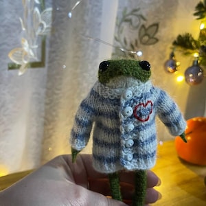 Könnte beinhalten: Handgefertigte, gestrickte Froschfigur mit einem blau-weiß gestreiften Pullover mit rotem Herzdetail. Der Frosch hat einen grünen Kopf und Beine sowie schwarze Augen. Der Pullover hat weiße Knöpfe.