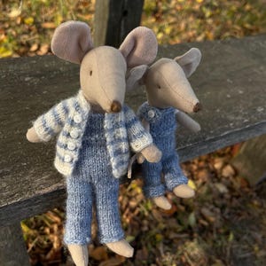 May include: Two plush toy mice are seated on a wooden bench. The larger mouse wears a blue and white striped jacket and blue pants. The smaller mouse wears blue overalls. Both have tan ears and noses.