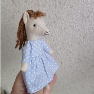 May include: A handmade doll in the shape of a horse. The horse has a cream-colored head with brown yarn hair and a light blue dress with white polka dots. The doll is made of fabric and has small black eyes.
