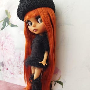 May include: A doll with long, vibrant orange hair, wearing a black knitted beret, dress, and boots. The doll has large eyes and is posed against a floral background. The dress features a fringed hem.