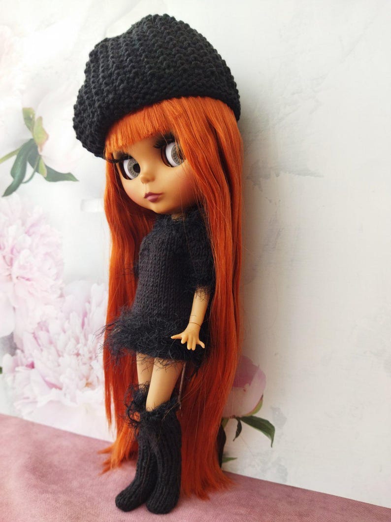 May include: A doll with long, vibrant orange hair, wearing a black knitted beret, dress, and boots. The doll has large eyes and is posed against a floral background. The dress features a fringed hem.