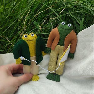 Könnte beinhalten: Zwei handgefertigte Filz-Froschpuppen mit Wackelaugen, eine gelb und eine grün, bekleidet mit Miniaturjacken und Hosen. Die Puppen sind durch eine dünne weiße Schnur mit kleinen weißen ohrhörerähnlichen Aufsätzen miteinander verbunden.