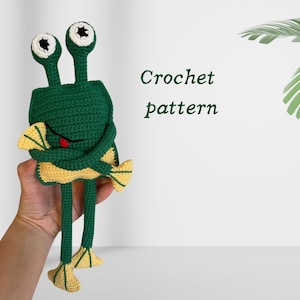 May include: Crochet pattern for a green frog holding a yellow fish. The frog has large, black eyes and a red tongue.