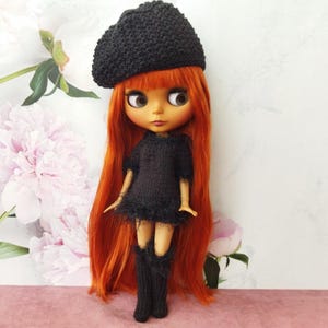 May include: A doll with long, vibrant red hair, wearing a black knitted beret, dress, and leg warmers. The doll has large eyes and is posed against a backdrop of soft pink and white floral patterns.