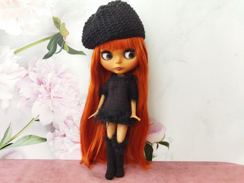 May include: A doll with long, vibrant red hair, wearing a black knitted beret, dress, and leg warmers. The doll has large eyes and is posed against a backdrop of soft pink and white floral patterns.