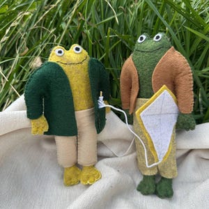 Könnte beinhalten: Zwei Froschpuppen aus Filz stehen nebeneinander. Der linke Frosch ist gelb mit einer dunkelgrünen Jacke und beigen Hosen. Der rechte Frosch ist grün mit einer braunen Jacke, beigen Hosen und hält einen weißen Drachen mit gelbem Rand an einer Schnur.