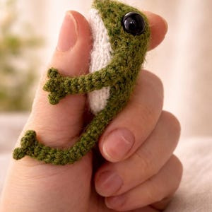 May include: A handmade green crochet frog with a white belly and large black eyes. The frog is small, with detailed legs and arms, and is held in a hand. The frog is a whimsical, handcrafted item.
