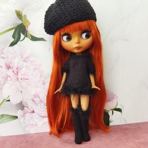 May include: A doll with long, vibrant red hair, wearing a black knitted beret, dress, and knee-high socks. The doll has large, expressive eyes and is posed against a backdrop of soft pink and white flowers.