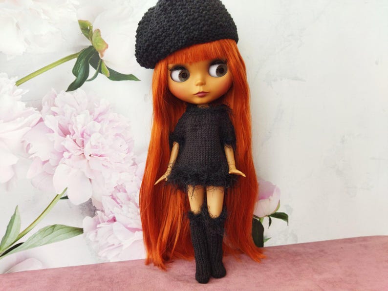 May include: A doll with long, vibrant red hair, wearing a black knitted beret, dress, and knee-high socks. The doll has large, expressive eyes and is posed against a backdrop of soft pink and white flowers.