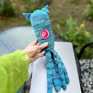 Op de afbeelding: Een handgemaakt, blauw en teal gehaakt inktvis speelgoed met een roze knoop en gestreepte details. De inktvis heeft twee kleine vinnen en lange tentakels. Het speelgoed wordt in de hand van een persoon gehouden, wat het vakmanschap en het grillige ontwerp laat zien.