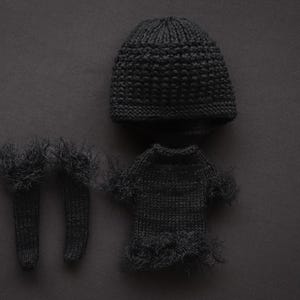 May include: A dark gray knitted baby clothing set, including a beanie, a sweater, and a pair of socks. The sweater and socks have a fringed trim. The set is displayed on a dark gray background.