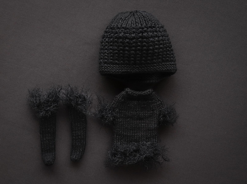 May include: A dark gray knitted baby clothing set, including a beanie, a sweater, and a pair of socks. The sweater and socks have a fringed trim. The set is displayed on a dark gray background.