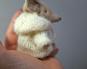 May include: A miniature stuffed mouse with a beige body and a cream-colored knitted coat. The mouse has a long, pointed nose and large ears. The toy is small enough to fit in the palm of a hand.