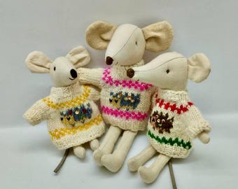 May include: Three plush mice toys with cream-colored bodies and large ears. Each mouse wears a knitted sweater with colorful patterns. The sweaters feature designs in yellow, pink, and red, with green and brown accents. The mice are sitting.