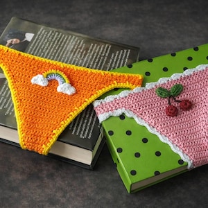 Può includere: Due segnalibri all'uncinetto a forma di biancheria intima, uno arancione con un arcobaleno e l'altro rosa con ciliegie. Il segnalibro arancione ha bordi gialli e quello rosa ha bordi bianchi. Sono posizionati sopra i libri.