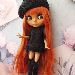 May include: A doll with long, vibrant orange hair and large eyes, wearing a black knitted beret, dress with a feathered trim, and knee-high socks. The doll is posed against a floral and pastel background.