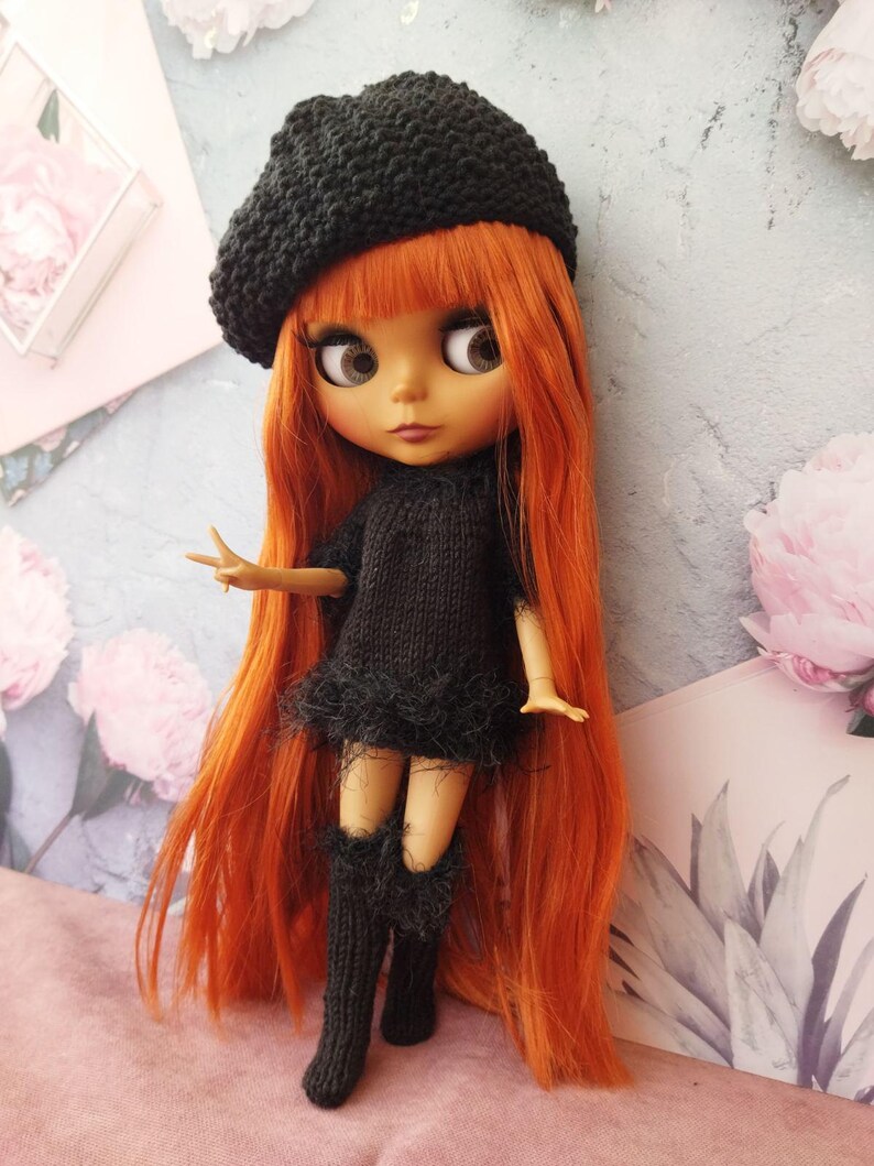 May include: A doll with long, vibrant orange hair and large eyes, wearing a black knitted beret, dress with a feathered trim, and knee-high socks. The doll is posed against a floral and pastel background.
