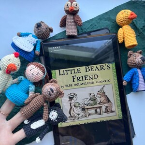 Pu&ograve; includere: Una collezione di marionette a dito all'uncinetto fatte a mano, tra cui un orso, un gufo, un'anatra e una ragazza, disposte attorno a un tablet che mostra il libro "Little Bear's Friend". Le marionette sono in vari colori, con dettagliati tratti del viso e abiti.