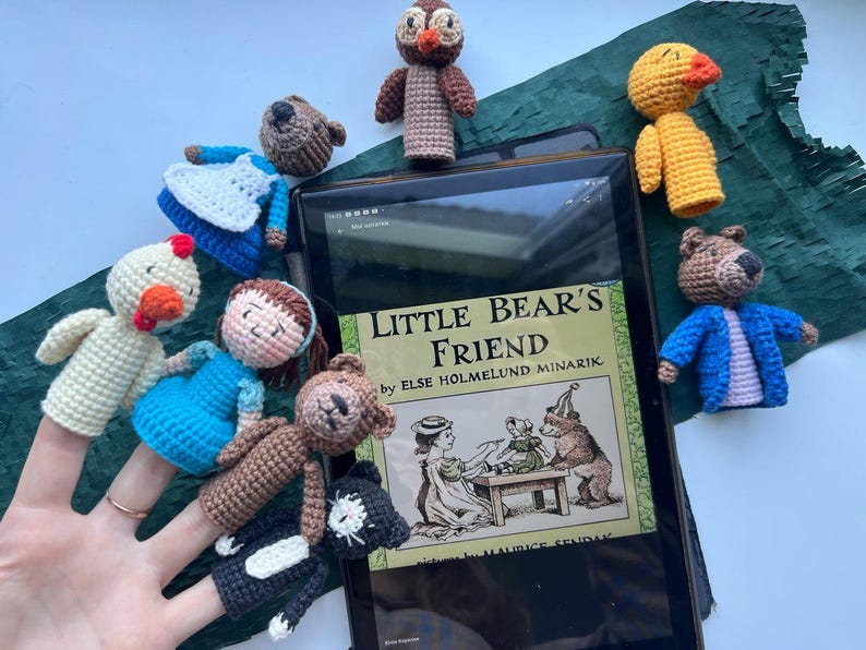 Pu&ograve; includere: Una collezione di marionette a dito all'uncinetto fatte a mano, tra cui un orso, un gufo, un'anatra e una ragazza, disposte attorno a un tablet che mostra il libro "Little Bear's Friend". Le marionette sono in vari colori, con dettagliati tratti del viso e abiti.