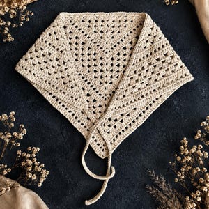May include: A beige triangular crochet headscarf featuring an openwork granny square pattern with a decorative border and two thin crocheted ties extending from the bottom corners to allow for secure fastening around the head or neck.