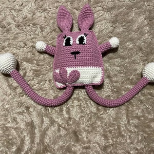 May include: A pink crocheted bunny with long legs and white accents. The bunny has large black eyes and a small black nose.