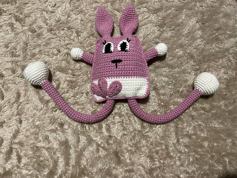 May include: A pink crocheted bunny with long legs and white accents. The bunny has large black eyes and a small black nose.