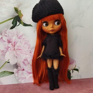 May include: A doll with long, vibrant red hair, wearing a black knitted beret, dress, and knee-high socks. The doll has large eyes and is posed against a backdrop of pink and white flowers.