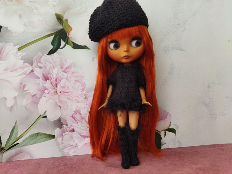 May include: A doll with long, vibrant red hair, wearing a black knitted beret, dress, and knee-high socks. The doll has large eyes and is posed against a backdrop of pink and white flowers.