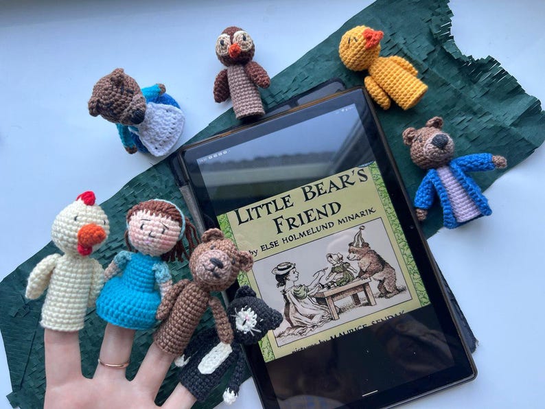 Pu&ograve; includere: Una collezione di marionette a dito fatte a mano, tra cui un orso, un gufo, un'anatra e una bambina, disposte attorno a un tablet che mostra la copertina del libro "Little Bear's Friend". Le marionette sono di vari colori, con illustrazioni sulla copertina.