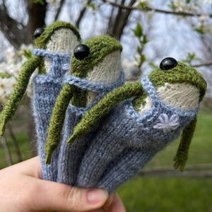 May include: Three handmade knitted frog dolls. Each frog has a green head, black eyes, and green arms. They are wearing blue overalls with a white collar and a small white flower detail. The background is blurred with white flowers.