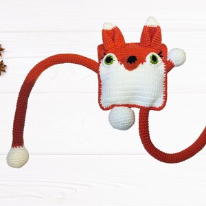 May include: A handmade crochet fox toy with a red body, white face, and green eyes. The fox has long, flexible arms and a tail, each ending in a white ball. The toy is designed with a whimsical, cartoonish style.