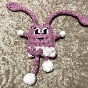 May include: A pink crocheted bunny toy with white accents. The bunny has large eyes, a small nose, and a heart-shaped tail.