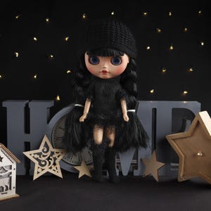 May include: A doll with blue eyes and black hair, wearing a black dress, hat, and boots. The doll is posed in front of a gray "HOME" sign, a clock, and wooden star decorations. A small wooden house with a deer cutout is also present.