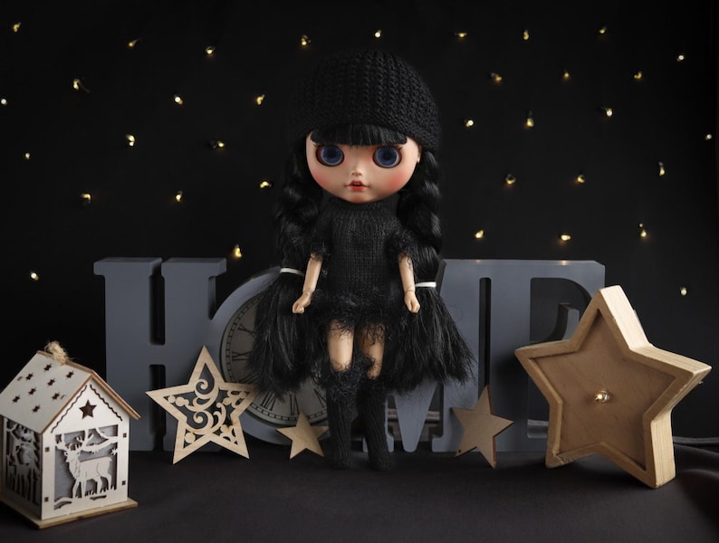 May include: A doll with blue eyes and black hair, wearing a black dress, hat, and boots. The doll is posed in front of a gray "HOME" sign, a clock, and wooden star decorations. A small wooden house with a deer cutout is also present.