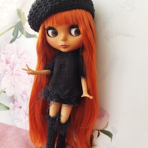 May include: A doll with long, vibrant orange hair, wearing a black knitted beret, dress, and knee-high boots. The dress has a feathered trim. The doll has large brown eyes and a fair complexion. The background features a floral pattern.