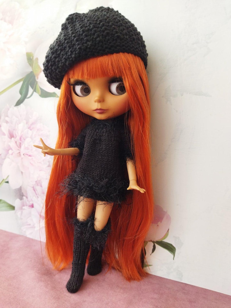 May include: A doll with long, vibrant orange hair, wearing a black knitted beret, dress, and knee-high boots. The dress has a feathered trim. The doll has large brown eyes and a fair complexion. The background features a floral pattern.