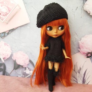 May include: A doll with long, vibrant orange hair, wearing a black beret, dress, and knee-high socks. The doll is posed against a backdrop with floral elements and a light blue and pink color scheme. The doll's dress has a fluffy trim.