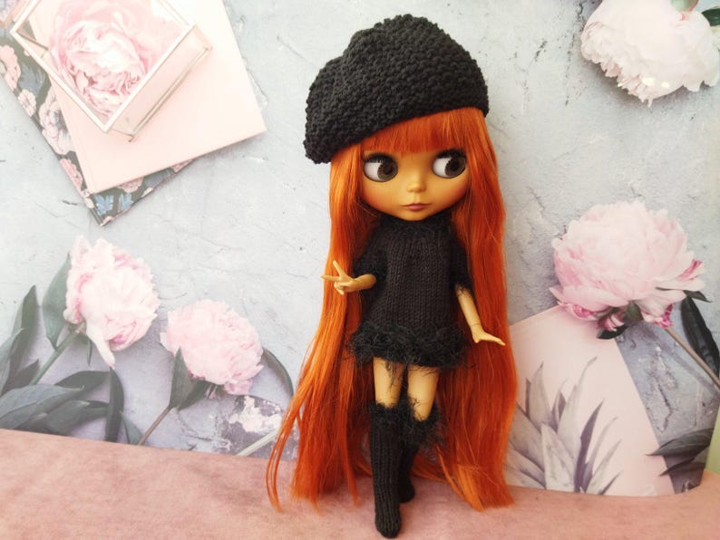 May include: A doll with long, vibrant orange hair, wearing a black beret, dress, and knee-high socks. The doll is posed against a backdrop with floral elements and a light blue and pink color scheme. The doll's dress has a fluffy trim.