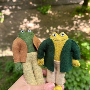 Könnte beinhalten: Zwei handgefertigte Froschpuppen aus Filz. Der linke Frosch ist dunkelgrün mit einer beigen Jacke und hellgelber Hose. Der rechte Frosch ist gelb mit einer dunkelgrünen Jacke und beiger Hose. Beide haben aufgestickte Münder und kleine weiße Augen.