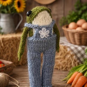 Könnte beinhalten: Handgestrickte Froschpuppe mit grünen Armen und Kopf, cremefarbenem Gesicht und blauer Latzhose mit weißer Blume. Der Hintergrund zeigt Sonnenblumen, Heuballen und Karotten.
