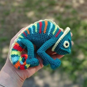 May include: A crocheted chameleon toy in teal and blue with a rainbow tail. The handmade reptile has a textured body and is held in a hand. The chameleon's head is a darker teal with a cream-colored trim. A unique, handcrafted item.