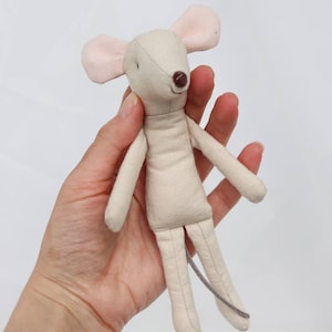 Pode incluir: Uma boneca de rato de pelúcia feita à mão em uma paleta de cores neutras. A boneca apresenta um corpo bege claro, orelhas rosa e um nariz marrom. Possui braços e pernas longos e uma cauda cinza. A boneca é feita de tecido macio.