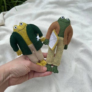Könnte beinhalten: Zwei handgefertigte Froschpuppen aus Filz. Der linke Frosch hat einen gelben Körper, eine dunkelgrüne Jacke und eine beige Hose. Der rechte Frosch hat einen dunkelgrünen Körper, eine braune Jacke, eine beige Hose und hält ein weißes dreieckiges Objekt.