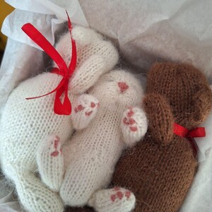 May include: Two knitted soft toys, a white bunny and a brown bear, presented in white tissue paper. The bunny has pink paw pads and a red ribbon. The bear also has a red ribbon. The toys are handmade.