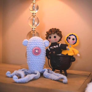 May include: Crocheted dolls and an octopus toy are displayed on a wooden surface. The dolls include a figure in a yellow coat and another in a black sweater. The octopus is light blue with a pink button.