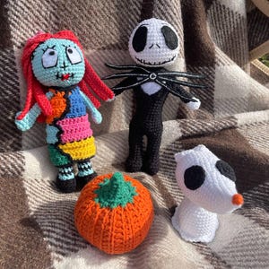 May include: A collection of crocheted figures including Sally, Jack Skellington, Zero, and an orange pumpkin. Sally has red hair and a patchwork dress. Jack is in a black suit. Zero is white with black spots. The pumpkin is orange.